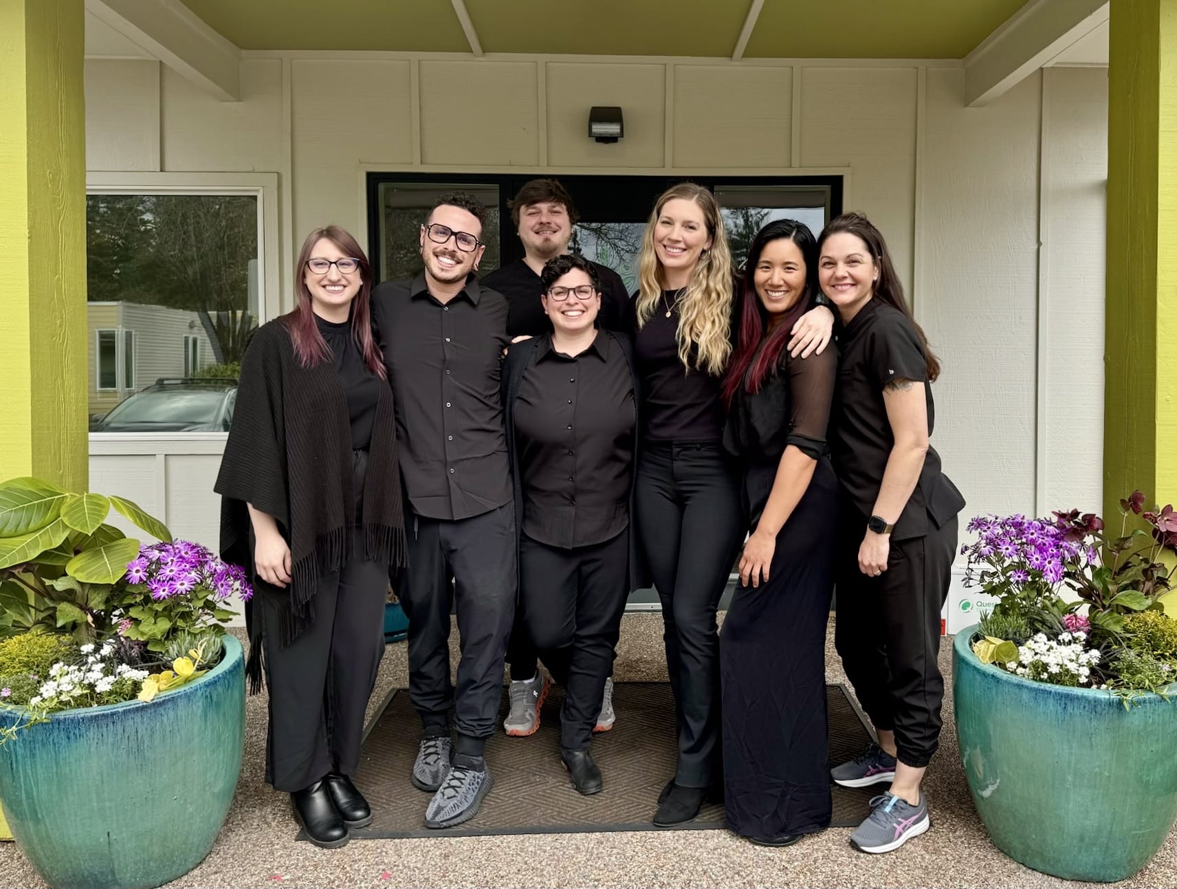 <br />
ChatGPT<br />
The image shows a group of seven people posing together in front of a light-colored house entrance, all dressed in stylish black attire. They are smiling broadly, displaying a warm, friendly demeanor. The group is diverse, consisting of three men and four women. Behind them are vibrant flower pots filled with purple and white flowers, adding a lively touch to the setting. The yellow-painted house door and greenery around the porch provide a pleasant, welcoming atmosphere.