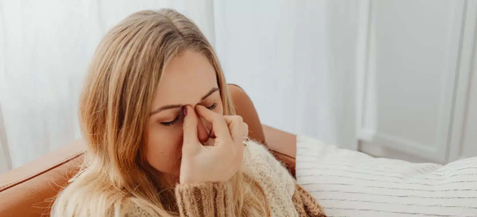 Blonde woman in cozy knit sweater pinching nose bridge, experiencing sinus pressure from MCAS and histamine intolerance