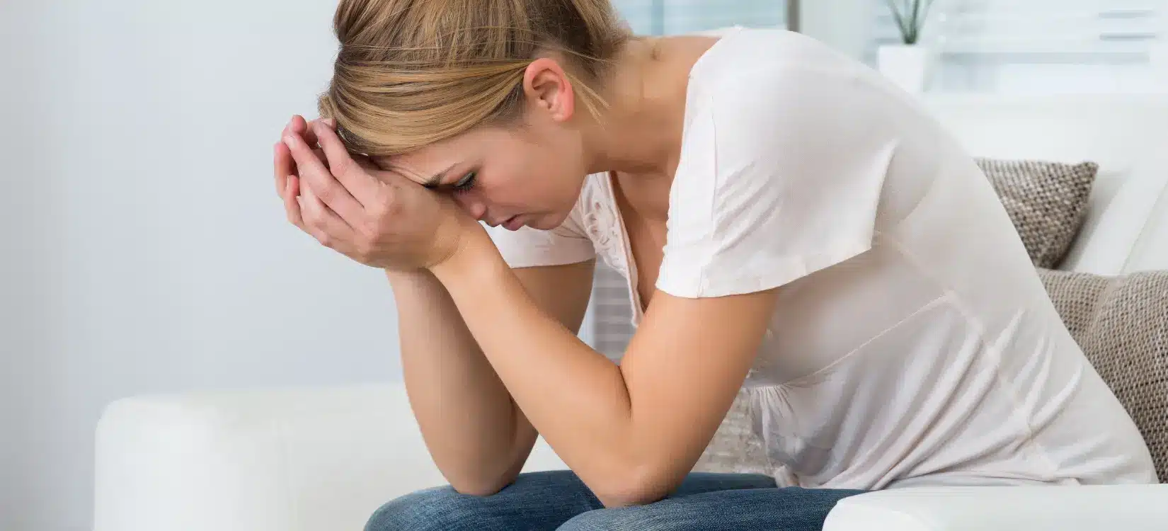 Distressed woman sitting on couch with head in hands, experiencing MCAS and POTS symptoms like fatigue and headache